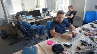 Ákos cikkírást imitál a fotó kedvéért, mögötte Anti és Elvisz lazsálnak, a háttérben Nyegleó feje búbja lóg ki a monitor mögül