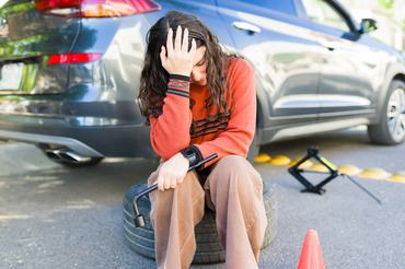 Teenage girl sitting on spare tire holding lug wrench with broken-down car and warning cone, experiencing frustration and stress