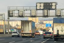 A section of the M0 motorway in Budapest showing a moderate flow of traffic, including cars and trucks. The image features overhead signage structures and an urban cityscape in the background, indicating the motorway's proximity to the city. The road surface is marked with clear lines and arrows, guiding traffic.