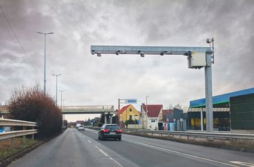 This image depicts a rural highway scene near Budapest, Hungary, with a metal structure spanning across the road. Mounted on this structure is a camera system used for traffic enforcement, specifically for monitoring vehicle speeds on the M5 motorway. The image shows a grey, overcast sky and a road with some light traffic. The camera system is designed to capture images of vehicles exceeding the posted speed limits.