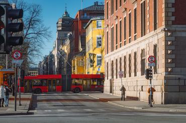 The image shows a red city bus driving through a central street in Oslo, Norway on a clear winter morning with soft sunlight illuminating colorful historic buildings, classic European architecture, pedestrians, traffic signs, and urban details reflecting everyday city life in the Norwegian capital