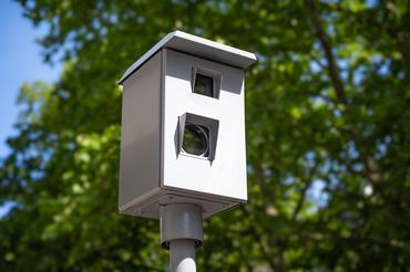 Traffic speed camera mounted on a pole outdoors with green trees and blue sky in the background